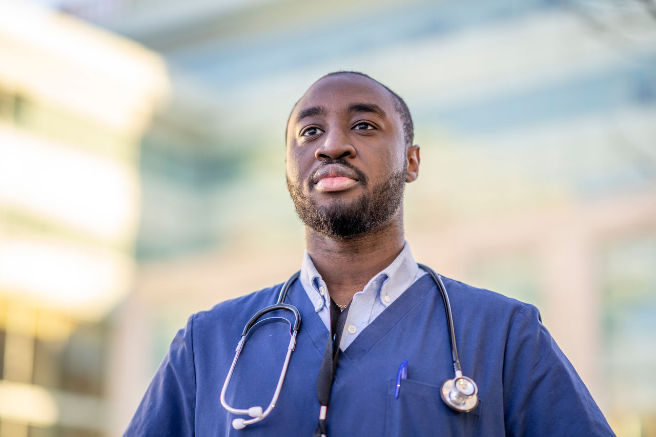 A portrait of a male nurse of African descent standing outside a hospital building. He is wearing blue medical scrubs and is looking away from the camera.